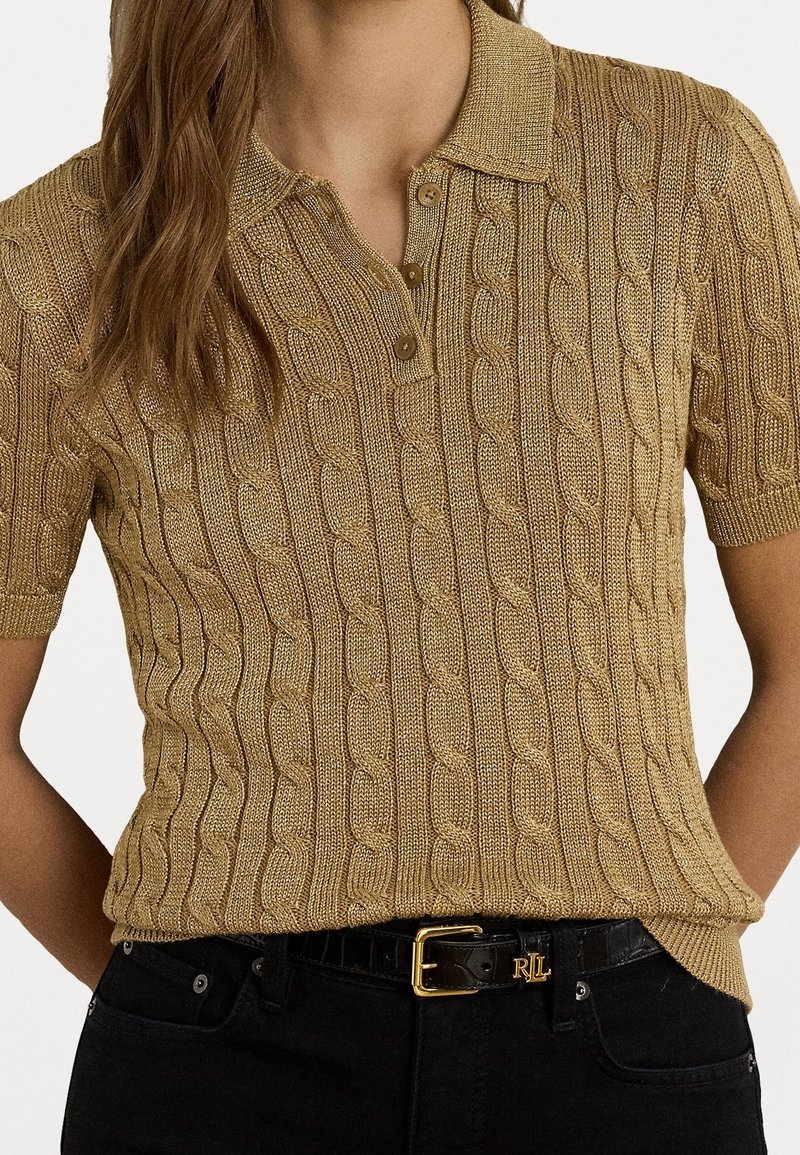 Femme portant un polo à manches courtes beige en maille torsadée, rentré dans un pantalon noir avec une ceinture noire avec boucle dorée.