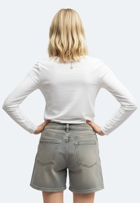 Persona bionda con capelli alla spalla si trova in piedi con le mani sui fianchi, indossando una camicia bianca a maniche lunghe e pantaloni corti in denim grigio a vita alta.