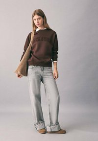Giovane donna che indossa un felpa marrone, jeans a gamba larga di un grigio chiaro, scarpe beige e porta una grande borsa a tracolla beige, in piedi contro uno sfondo neutro.