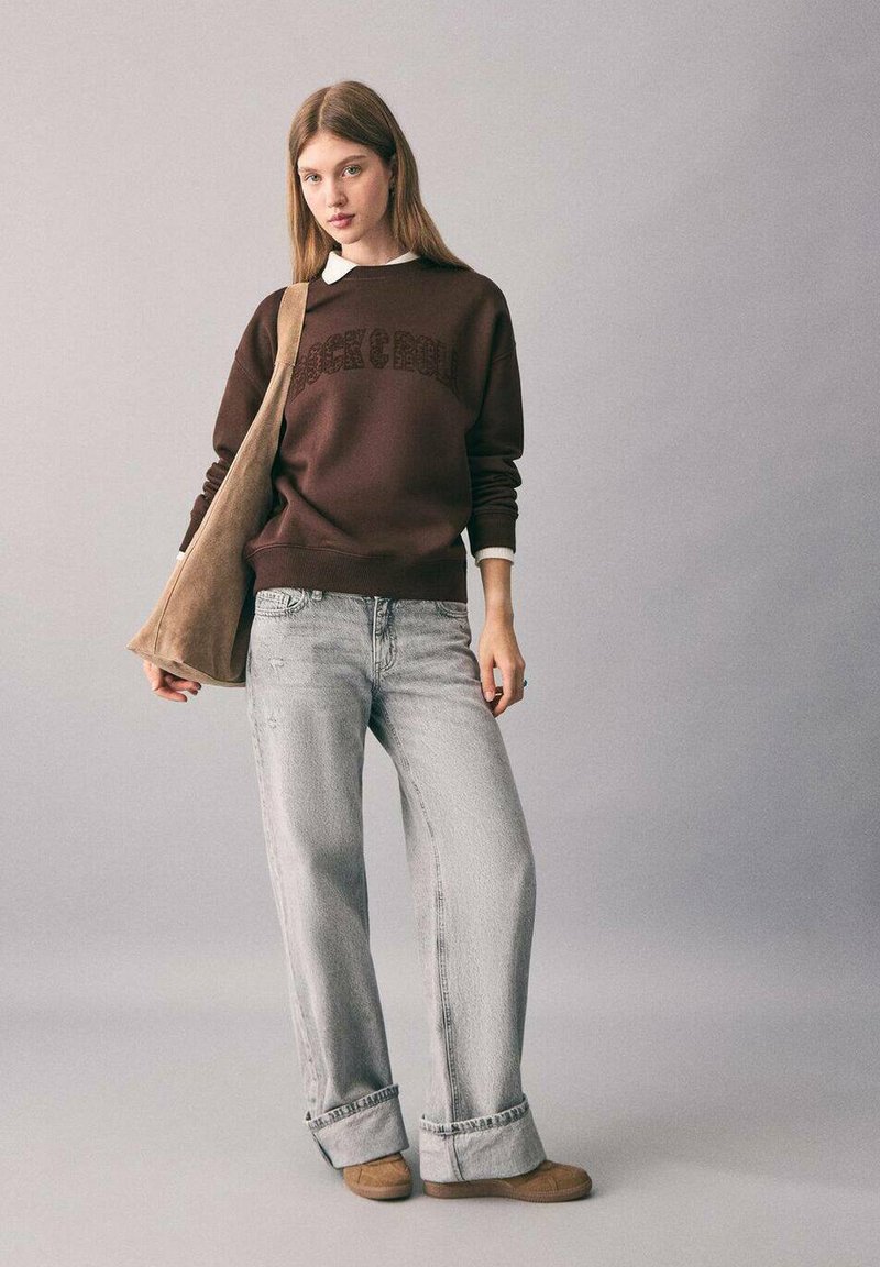 Giovane donna che indossa un felpa marrone, jeans a gamba larga di un grigio chiaro, scarpe beige e porta una grande borsa a tracolla beige, in piedi contro uno sfondo neutro.