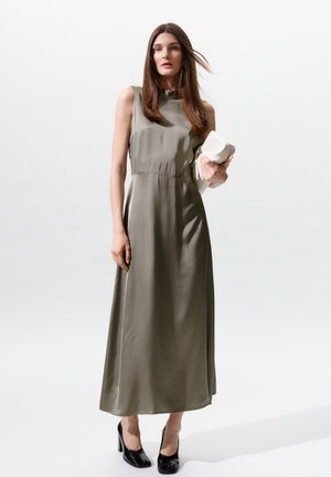 Femme aux longs cheveux bruns portant une robe maxi sans manches vert olive, tenant une pochette blanche, debout sur un fond blanc.