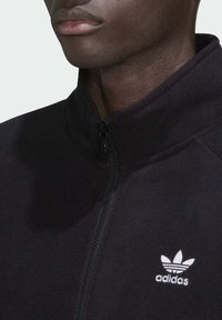 Must musta tõmblukkidega jakk kõrge kaelusega, millel on vasakul rindkere piirkonnas valge Adidas logo ja tekstuurne kangas.