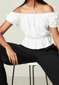 Haut blanc décolleté avec des manches froncées et une taille cintrée, associé à un pantalon noir texturé, assis sur une chaise blanche.