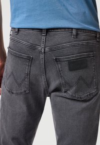 Jeans de mezclilla gris con costuras en contraste, un diseño de bolsillo único con bordes curvados y un parche de logo de Wrangler en negro en la parte trasera.