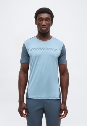 Bărbat stând cu fața spre înainte îmbrăcat într-un tricou sport ușor albastru deschis cu mânecă scurtă cu logo-ul "DYNAFIT" și pantaloni albaștri asortați pe fundal alb.