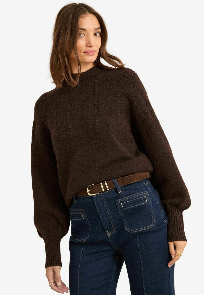 Brauner Strickpullover mit Rundhals, strukturiertem Muster und gerippten Bündchen, kombiniert mit blauen Jeans und einem braunen Gürtel mit goldfarbenen Details.