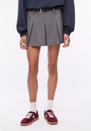 Persona che indossa una gonna plissettata corta grigia con cintura nera, felpa blu navy, calzini bianchi e sneakers bordeaux, in piedi davanti a uno sfondo semplice.