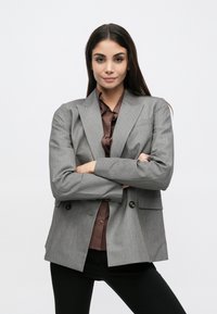 Blazer gris à finition texturée, deux boutons et poches avant, porté sur une chemise marron boutonnée et un pantalon noir.