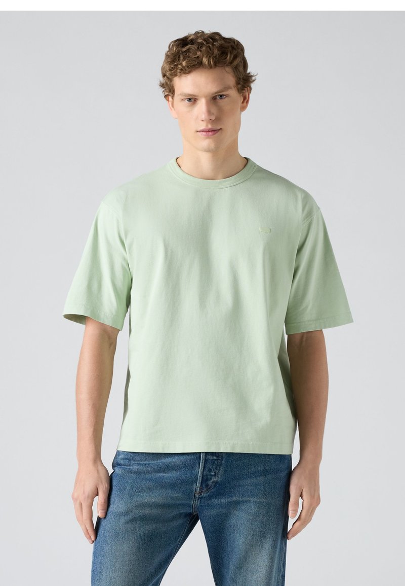 Jeune homme aux cheveux bouclés portant un t-shirt à manches courtes vert clair et un jean bleu, debout devant un fond gris clair uni.