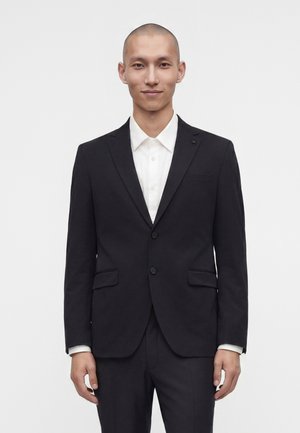 Bărbat cu capul ras, purtând un sacou și pantaloni negri de costum, cu o cămașă albă, stând în fața unui fundal alb simplu.