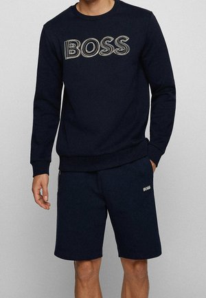 Homme portant un sweat-shirt bleu marine avec le texte "BOSS" et un short bleu marine assorti avec un petit logo "BOSS" sur la cuisse gauche.