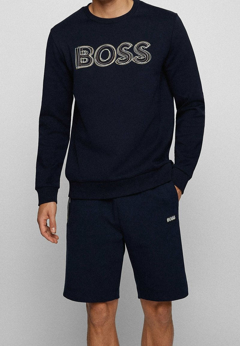 Homme portant un sweat-shirt bleu marine avec le texte "BOSS" et un short bleu marine assorti avec un petit logo "BOSS" sur la cuisse gauche.
