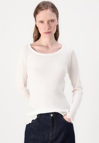 Valkoinen ribbed pitkähiha-olemus, jossa on leveä kaula-aukko ja aaltoilevat hihansuut, yhdistettynä tummiin farkkuihin. Kangas on pehmeän tuntuista.