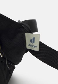 Bolsa de tela negra con una textura suave, una etiqueta de logotipo prominente y una correa resistente. Detalles de cremallera visibles en el lateral.
