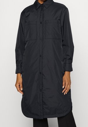 Abrigo largo negro abotonado con cuello, dos bolsillos en el pecho y mangas largas, llevado por una persona con las manos relajadas a los costados.