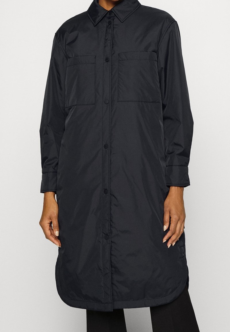Long manteau noir à boutons avec col, deux poches poitrine et manches longues porté par une personne les mains détendues le long du corps.