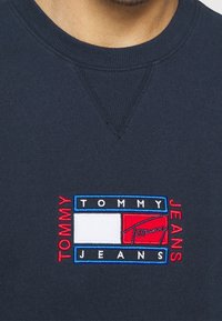 Marinblå sweatshirt med en rektangulär broderad logotyp som har "TOMMY" i rött och "JEANS" i vitt, med en vit och röd färgblockdesign.