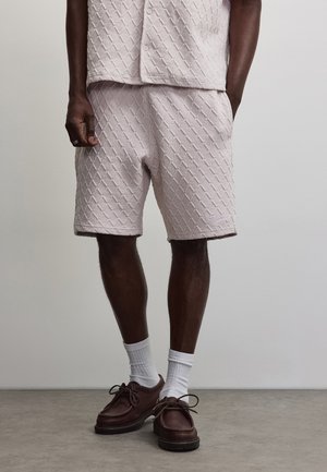Persona con pantalones cortos texturizados de color rosa claro y camisa a juego, calcetines blancos y zapatos marrones con cordones, de pie sobre un suelo gris.