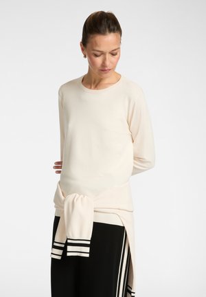 Donna con i capelli raccolti, che indossa una maglia a maniche lunghe color crema con un maglione abbinato annodato in vita e pantaloni neri con righe bianche.