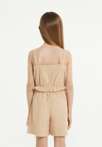 Beiger Leinen-Overall mit verstellbaren Trägern, tailliertem Schnitt und Rüschendetails. Glatte Textur, kurze Beine und ein einfaches, lässiges Design.
