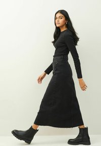 Mujer caminando con una camiseta negra de manga larga, falda vaquera negra de cintura alta y botas negras hasta el tobillo sobre un fondo claro y liso.