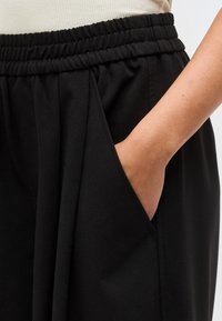 Mâna unei persoane odihnindu-se în buzunarul lateral al pantalonilor negri cu talie elastică, deasupra fiind vizibil un top alb cu model reiat.