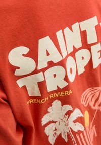 Camiseta de algodón roja con un atrevido texto blanco que dice "SAINT TROPEZ" y "FRENCH RIVIERA" debajo, acompañado de gráficos de palmeras. Textura suave.