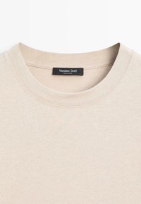 T-shirt girocollo in cotone beige con vestibilità regolare, caratterizzata da un'etichetta cucita sul colletto. Texture liscia e design semplice.