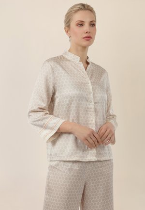 Frau trägt ein cremefarbenes Satin-Pyjama-Set mit einem sechseckigen Muster, steht mit verschränkten Händen vor einem beigefarbenen Hintergrund.