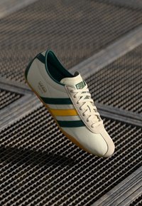 Adidași albi cu dungi verzi și galbene, realizați din material sintetic neted. Prezintă un vârf rotund și o talpă plată din cauciuc.