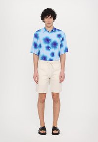 Cămașă cu mânecă scurtă, de culoare albastră, cu model tie-dye albastru, asortată cu pantaloni scurți bej și sandale negre cu talpă plată. Design minimalist, fit casual.