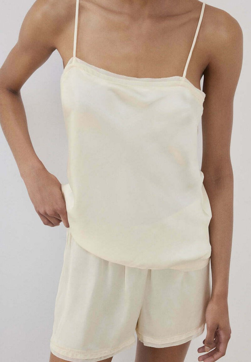 Personne portant un caraco ample en satin couleur crème et un short assorti avec des bretelles fines, debout devant un fond clair uni.