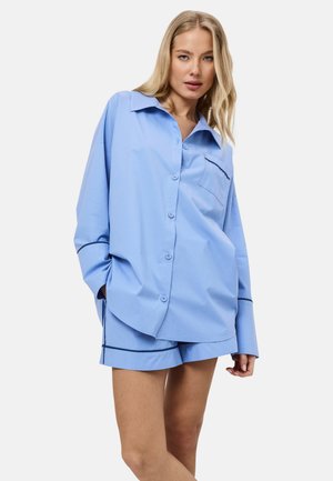 Blond meisje draagt lichtblauw oversized overhemd met knopen en bijpassende short met donkere rand, staand tegen een witte achtergrond.