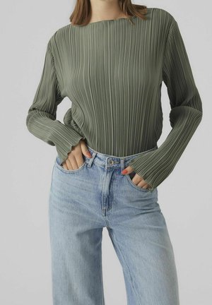 Person iført en ribbet olivengrøn langærmet top og lyseblå high-waisted jeans med hænderne i jeanslommerne.