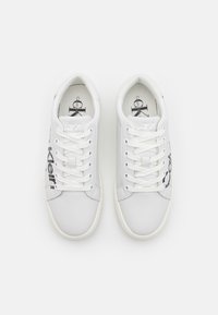 Ett par vita Calvin Klein snörskor med svart logotyp på sidorna, visade ovanifrån på en vit bakgrund.