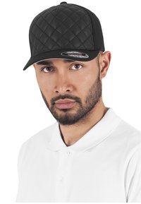 Cappello trapuntato nero con corona piatta e visiera rigida, indossato da un individuo in una polo bianca, che mette in mostra il materiale testurizzato e la forma.
