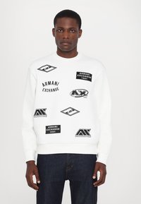 Biała bluza z różnymi czarnymi graficznymi naszywkami i logo, ściągacze przy mankietach i u dołu, okrągły dekolt, noszona z ciemnoniebieskimi dżinsami.