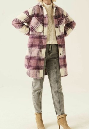 Vrouw die een lange roze-witte geruite jas draagt over een crèmekleurige kabelgebreide trui, grijze jeans en beige suède enkellaarsjes.