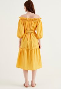 Off-shoulder gul klänning med puff�ärmar, åtsittande midja, volangdetaljer och ett lager-på-lager nederdel, tillverkad i lätt tyg.
