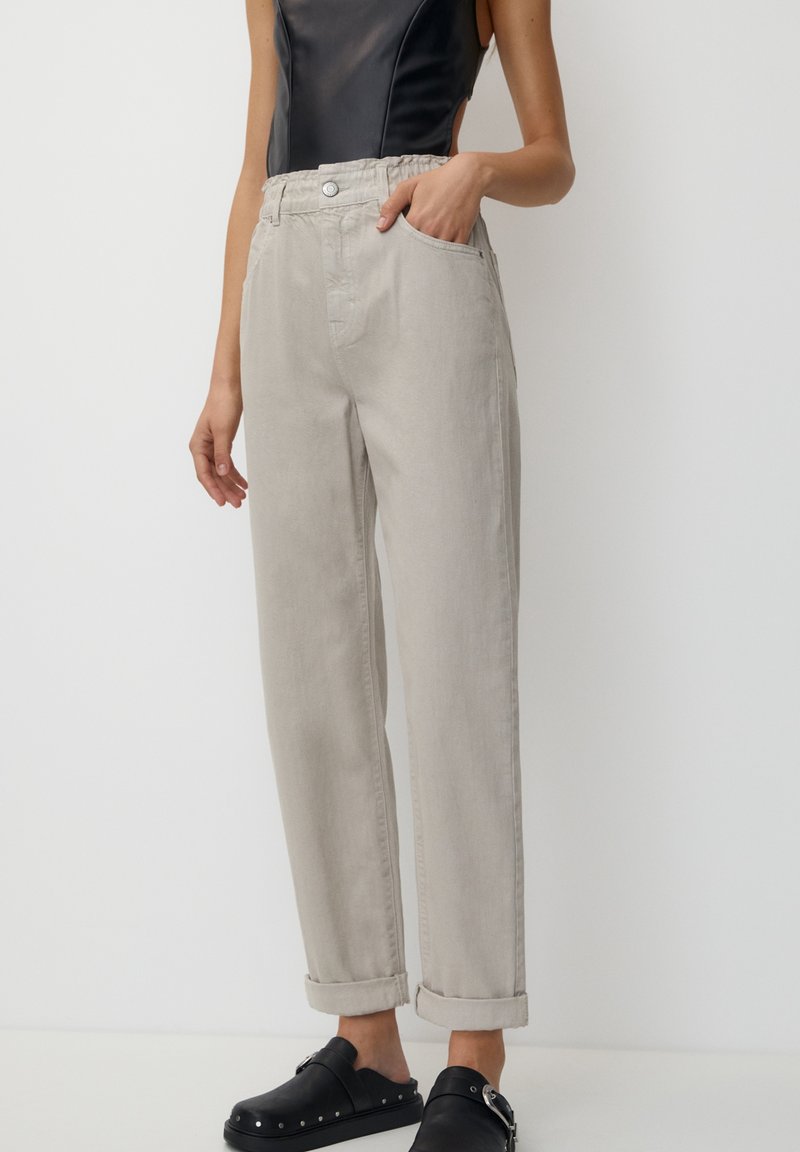 Pantalons gris clair taille haute avec une coupe décontractée, dotés de poches arrière et de revers roulés. Associés à des chaussures noires à enfiler.