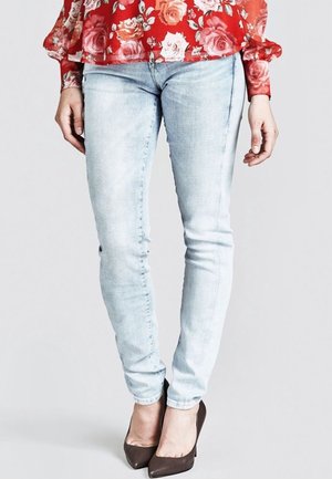 Femme portant un jean slim bleu clair, des talons pointus marron et un chemisier à manches longues à motif floral rouge, debout devant un fond uni.