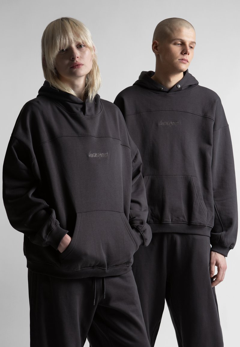 Twee jonge volwassenen in zwarte hoodies en zwarte broeken staan tegen een effen grijze achtergrond, met een serieuze blik naar voren gericht.