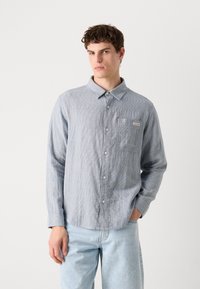 Világoskék csíkos hosszú ujjú ing gombos gallérral, elülső zsebbel és Calvin Klein logóval. Világoskék farmernadrággal viselve.