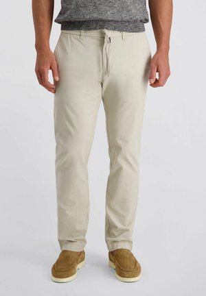 Mann trägt beige Kordelzughose, graues Hemd und braune Slipper, steht vor hellem, einfarbigem Hintergrund.