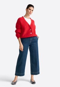 Mujer con un cárdigan rojo abotonado, camiseta blanca, pantalones vaqueros de pierna ancha azules y bailarinas negras, de pie con una mano en el bolsillo.