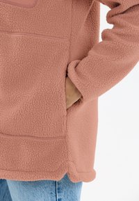 Weicher, strukturierten rosa Fleece-Jacke mit einer Seitentasche und abgerundetem Saum, getragen über lässigen blauen Denim-Jeans.