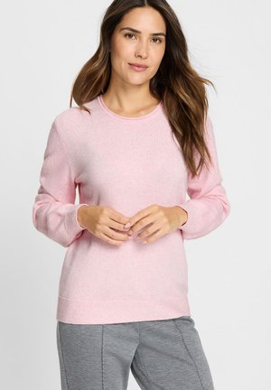 Frau mit langen braunen Haaren, die einen hellrosa Pullover und graue Hosen trägt, steht mit sanft vor der Taille verschränkten Händen.