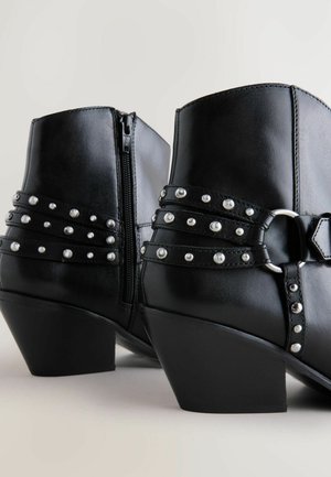 Fekete bőr bokacsizma cipzárral, ezüst tüskékkel és díszes pántokkal a középső részen, valamint tömbsarkal.