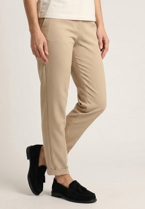 Person iført beige slim-fit bukser og sorte tasselloafers med opsmøgede kanter, der står mod en ensfarvet baggrund.