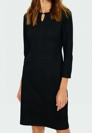 Schwarzes knielanges Kleid mit dezentem Karomuster, dreiviertel Ärmeln und Schlüssellochausschnitt mit Verschlussdetail, getragen von einer stehenden Frau.